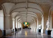 Vue du hall dans sa longueur, rythmé de lourds piliers de pierre sous une voûte cintrée parsemée de lumignons, à droite des statues, à gauche des guichets, au fond un lustre multicolore.
